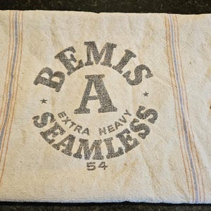 Puede incluir: Un saco de tela beige vintage con el texto "BEMIS EXTRA HEAVY SEAMLESS 54" en gris oscuro. El saco tiene rayas verticales azules y rojas y un aspecto desgastado con un desgarro.