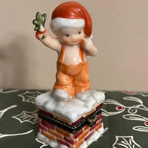 May include: A porcelain figurine of a cherubic child in orange overalls and a Santa hat, holding a sprig of holly. The figurine stands atop a chimney-shaped trinket box with a hinged lid, painted with brick detail and snow.