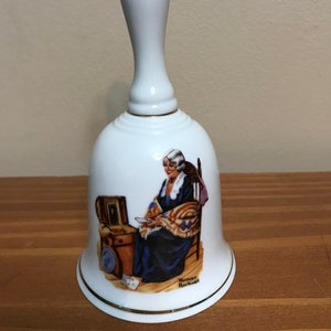 May include: White porcelain bell with a gold trim. The bell features a painting of a woman sitting in a chair, holding a letter. The painting is by Norman Rockwell.