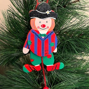 May include: A wooden clown Christmas ornament. The clown has a black hat with a white flower, a red nose, and a red and blue striped shirt. The clown's green pants and red shoes are visible. The ornament hangs from a red string.