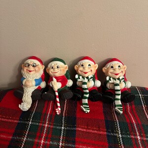 May include: Four Christmas stocking holders shaped like elves. The elves are wearing red and green hats, and holding candy canes. They are sitting on a red and green plaid surface.