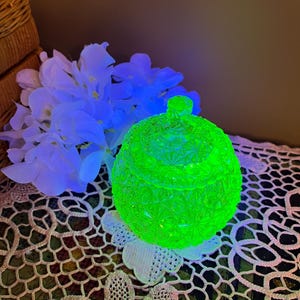 May include: A round, lidded glass jar featuring a pressed geometric star pattern that glows a vibrant neon green under ultraviolet light. The container has a small, circular knob on the lid and a textured, faceted surface throughout its exterior.