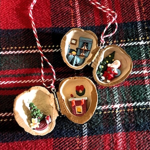 May include: Three Christmas ornaments crafted from walnut shells, each featuring a miniature holiday scene. One shows Santa Claus, another a Christmas tree, and the third a fireplace. They hang from red and white twine, set against a red and green plaid background.