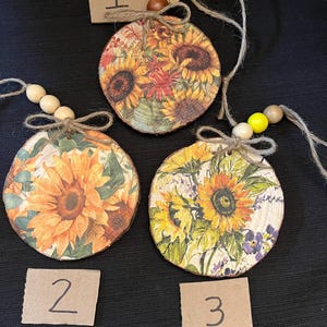 May include: Three round wooden ornaments with sunflower designs. Each ornament is decorated with a different sunflower pattern and has a twine loop with wooden beads for hanging. The ornaments are numbered 1, 2, and 3 on small cardboard tags.
