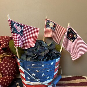 May include: A metal bucket painted with red, white, and blue stripes and white stars, containing dark blue artificial flowers and three small fabric flags on wooden sticks. The flags feature red and white gingham or striped patterns with decorative patches.