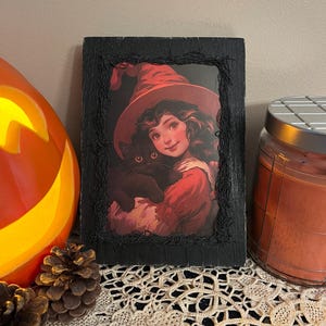 May include: A framed illustration of a young girl wearing a red witch hat and holding a black cat. The image is set against a dark background and has a vintage aesthetic.