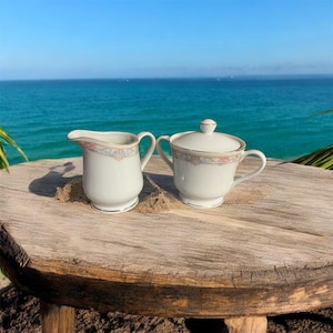 May include: A white porcelain creamer and sugar bowl set with a gold trim and a delicate seashell design. The set is displayed on a rustic wooden table with a beach and ocean background.
