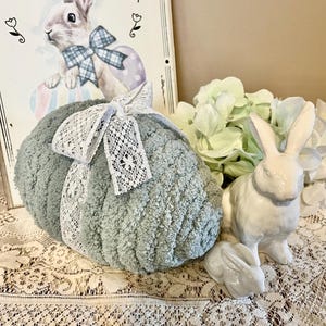 May include: A large, light blue, fuzzy Easter egg with a white lace ribbon. The egg is sitting on a white lace tablecloth with two white ceramic bunnies.