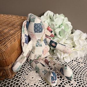 May include: A handmade patchwork bunny doll with a variety of floral and geometric patterned fabric pieces. The bunny is resting on a white crocheted doily, with a wicker basket and white and green artificial flowers in the background.