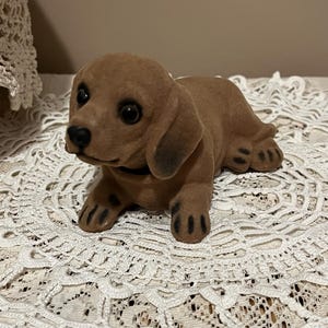 May include: A brown dachshund figurine with black accents on its ears, paws, and nose. The dog is lying down, with a black collar. The figurine is made of a soft, velvety material. The background is a white lace doily.