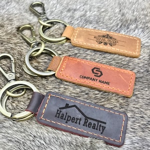 May include: Three leather keychains with metal key rings. The keychains are brown, tan, and black. The brown keychain has the text "Royal Hotel" on it. The tan keychain has the text "Company Name" on it. The black keychain has the text "Halpert Realty" on it.