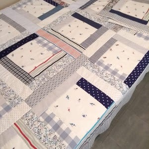 May include: A patchwork quilt featuring a mix of blue, white and grey fabrics. The quilt is made up of squares and rectangles of varying sizes and patterns. The quilt is displayed on a bed.