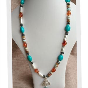 May include: A beaded necklace with turquoise, orange, and gray beads. The necklace has a silver pendant with a Celtic knot design and a turquoise bead. The necklace is displayed on a white bust.