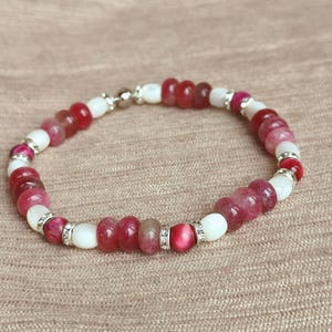 May include: A beaded bracelet featuring a mix of red, pink, and white beads. The bracelet has a silver clasp and small silver accents between some of the beads. The beads are round and vary in size.