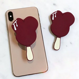 May include: A burgundy Mickey Mouse-shaped ice cream pop phone grip with a white stick and white drips on the top. The phone grip is attached to a gold iPhone.