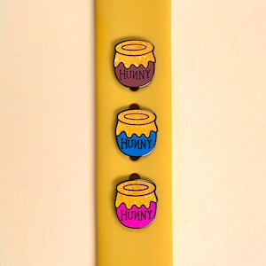 Puede incluir: Tres pines de esmalte con forma de tarro de miel con la palabra "Hunny" en ellos. Los pines son de diferentes colores: marrón, azul y rosa.