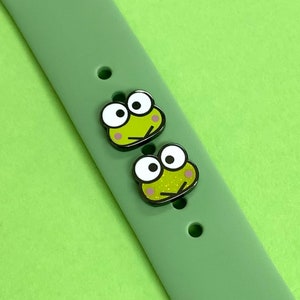 May include: Two green frog-shaped watch band charms with black outlines and pink cheeks. The charms are on a green silicone watch band.