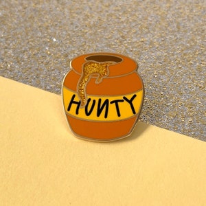 May include: A gold-colored enamel pin in the shape of a honey pot. The pot is orange with a yellow band and the word "HUNNY" in black. Gold glitter drips from the top of the pot.