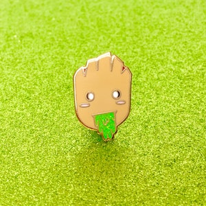 May include: A gold-colored enamel pin featuring a cartoon character with a green, gooey substance coming out of its mouth. The character has a brown face, white eyes, and a pink mouth.