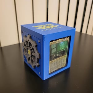 May include: A blue card storage box with the text "VAULT-TEC INDUSTRIES" in yellow. The box has a gear design on the side and a card visible through a window. The card features text and a green-tinted image.