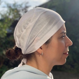May include: A white cotton head wrap tied in a knot at the back of the head. The wrap is worn over a light blue hooded sweatshirt.
