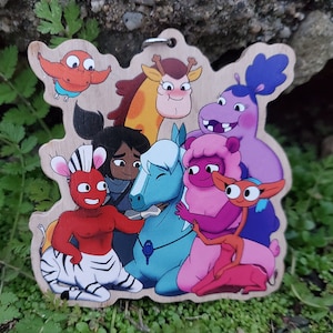 May include: Wooden cutout featuring a group of cartoon characters in various colors. The characters include a zebra-like figure, a horse, a giraffe, and others. The cutout has a natural wood grain finish and a metal ring for hanging.