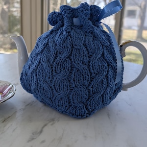 May include: A white porcelain teacup and saucer set with floral designs in pink, purple, and blue. A blue knitted tea cozy covers a white teapot. The cozy has a cable knit pattern and a blue ribbon. The set is on a white surface.