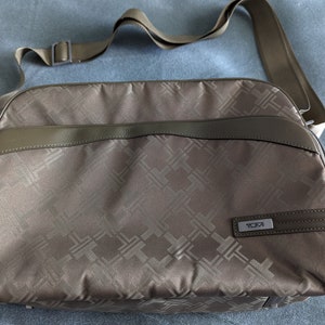 May include: A brown leather and fabric crossbody bag with a TUMI logo. The bag has a geometric pattern and a long adjustable strap.