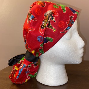 May include: Red surgical scrub hat with a pattern of various Marvel superheroes, including Thor, Hulk, Iron Man, and Spider-Man. The hat has a black tie and is tied in a knot at the back.