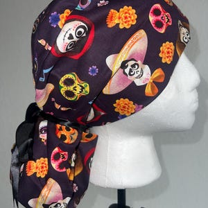 May include: A dark purple scrub cap featuring a repeating pattern of sugar skulls, marigold flowers, and characters from the movie "Coco." The cap has a black ribbon tie and is displayed on a white mannequin head.