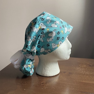 May include: A light blue scrub cap featuring a repeating pattern of Snoopy and Woodstock characters. The cap is tied in the back with a white ribbon. The design includes paw prints and flowers. The cap is displayed on a white mannequin head.