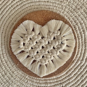 Può includere: Un sottobicchiere in macramé bianco a forma di cuore con retro in sughero. Il sottobicchiere è realizzato con un design a corda annodata e ha una frangia sui bordi.