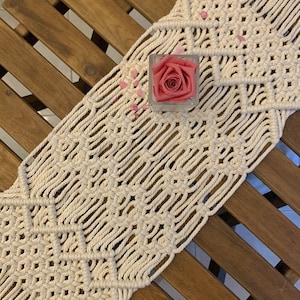 Può includere: Runner da tavolo in macramè color crema con motivo geometrico, posto su un tavolo di legno. Un piccolo cubo di vetro contiene una rosa rosa, con petali sparsi. Il design intrecciato aggiunge un tocco bohémien all'arredamento della tavola.