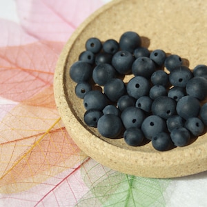 Può includere: Primo piano di una piccola ciotola piena di perline rotonde blu scuro. La ciotola è fatta di sughero e si trova su una superficie bianca con foglie rosa e arancioni.