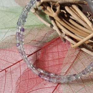 Peut inclure: Un collier délicat composé de petites perles carrées translucides dans des tons de violet, lavande et transparent. Le collier est drapé sur un fond de feuilles et de brindilles séchées, créant une esthétique naturelle. Le collier mesure environ 40 cm.
