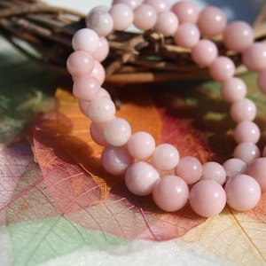 Peut inclure: Un bracelet en perles d'opale rose avec une finition chatoyante. Le bracelet est posé sur un fond de feuilles translucides colorées.