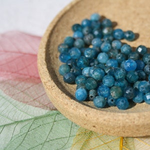Puede incluir: Un conjunto de cuentas facetadas de piedra preciosa azul-verdosa, probablemente apatito, llena un pequeño cuenco redondo de madera. Las cuentas tienen un aspecto cristalino y brillante. El cuenco se encuentra sobre una superficie con acentos coloridos en forma de hoja.