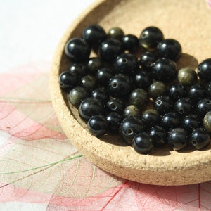 Può includere: Primo piano di una ciotola piena di perline di pietre preziose nere e dorate. Le perline sono rotonde e hanno una finitura liscia e lucida.