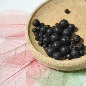 May include: A close-up of a small bowl filled with black lava rock beads. The beads are round and have a rough, porous texture. The bowl is made of cork and has a natural brown color.