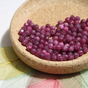 May include: A bowl of small, round, faceted, purple gemstone beads. The beads are in a shallow, cork-like bowl.