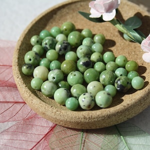 Peut inclure: Gros plan sur un bol rempli de perles de serpentine vertes. Les perles sont rondes et ont une finition lisse et polie. Le bol est en liège et a une couleur brune naturelle.