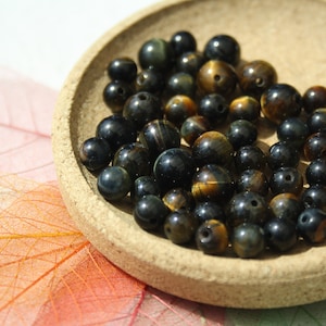 Op de afbeelding: Een close-up van een stapel donkerblauwe en gouden tijgeroog kralen. De kralen zijn rond en glad en hebben een metallic glans.