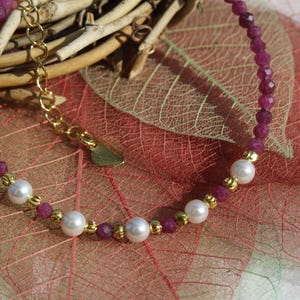 Peut inclure: Un bracelet délicat composé de petites perles rouge foncé facettées, entrecoupées de perles blanches crémeuses et d'éléments dorés. Le bracelet est posé sur un fond de feuilles et de brindilles rouges et vertes.