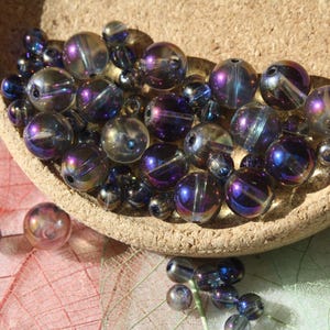 Peut inclure: Un assortiment de perles de verre iridescentes de différentes tailles, aux teintes violettes, bleues et argentées. Les perles sont contenues dans un petit bol en liège brun clair, avec quelques-unes éparpillées à l'extérieur. Les perles sont sur une feuille.