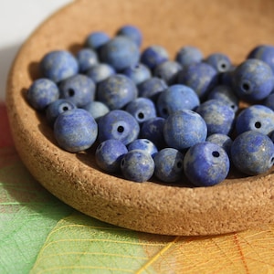 Può includere: Una collezione di perline blu in lapislazzuli dall'aspetto naturale e macchiettato, esposte in una ciotola rotonda di sughero. Le perline variano di dimensioni e hanno un foro. Lo sfondo presenta foglie traslucide colorate.