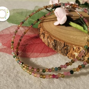 May include: A multi-strand necklace made of small, faceted, multi-coloured gemstones. The necklace is made of pink, green, and brown gemstones. The necklace is displayed on a wooden surface with a pink flower.