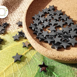 Peut inclure: Gros plan sur un tas de perles noires en forme d'étoile dans un petit bol en bois. Les perles sont éparpillées sur une surface en toile de jute brune et une feuille verte.