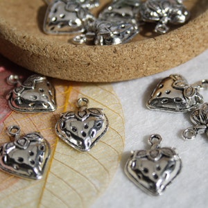 Puede incluir: Encantos de plata en forma de corazón con una superficie texturizada y un bucle para sujetar a joyas u otras manualidades. Los encantos están dispersos sobre una superficie blanca con una hoja en el fondo.