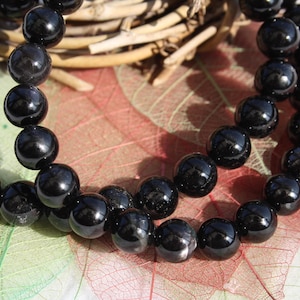 Puede incluir: Dos hilos de cuentas negras, brillantes y redondas, probablemente una pulsera o collar, se disponen sobre una superficie colorida y texturizada. Las cuentas reflejan la luz, creando reflejos. El fondo incluye hojas secas y ramitas.