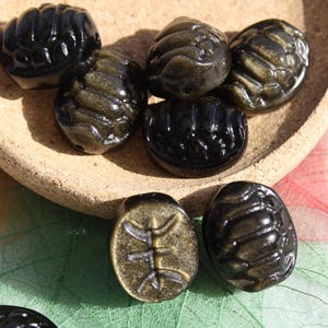 Puede incluir: Una colección de cuentas oscuras, ovaladas, con un acabado brillante, algunas con un brillo dorado, se exhiben en un cuenco poco profundo de color marrón claro. Una cuenta tiene un diseño tallado. Las cuentas están sobre un fondo colorido.